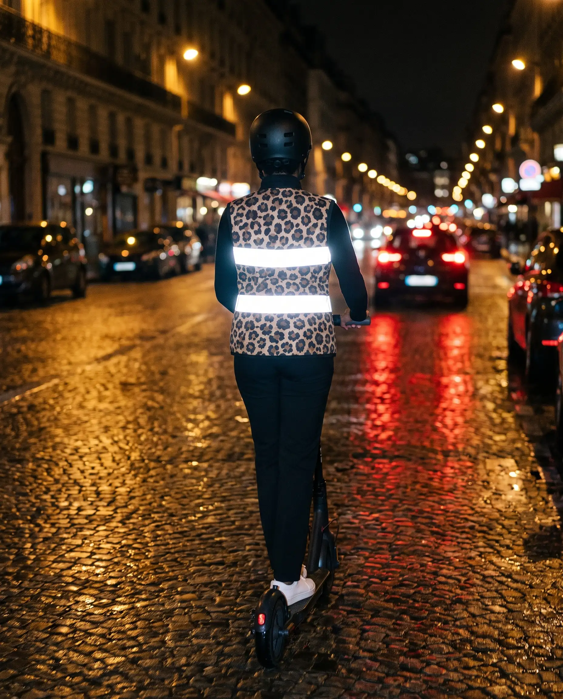 De dos, sur une trottinette, dans une rue pavée mouillée : les bandes du gilet brillent dans la nuit